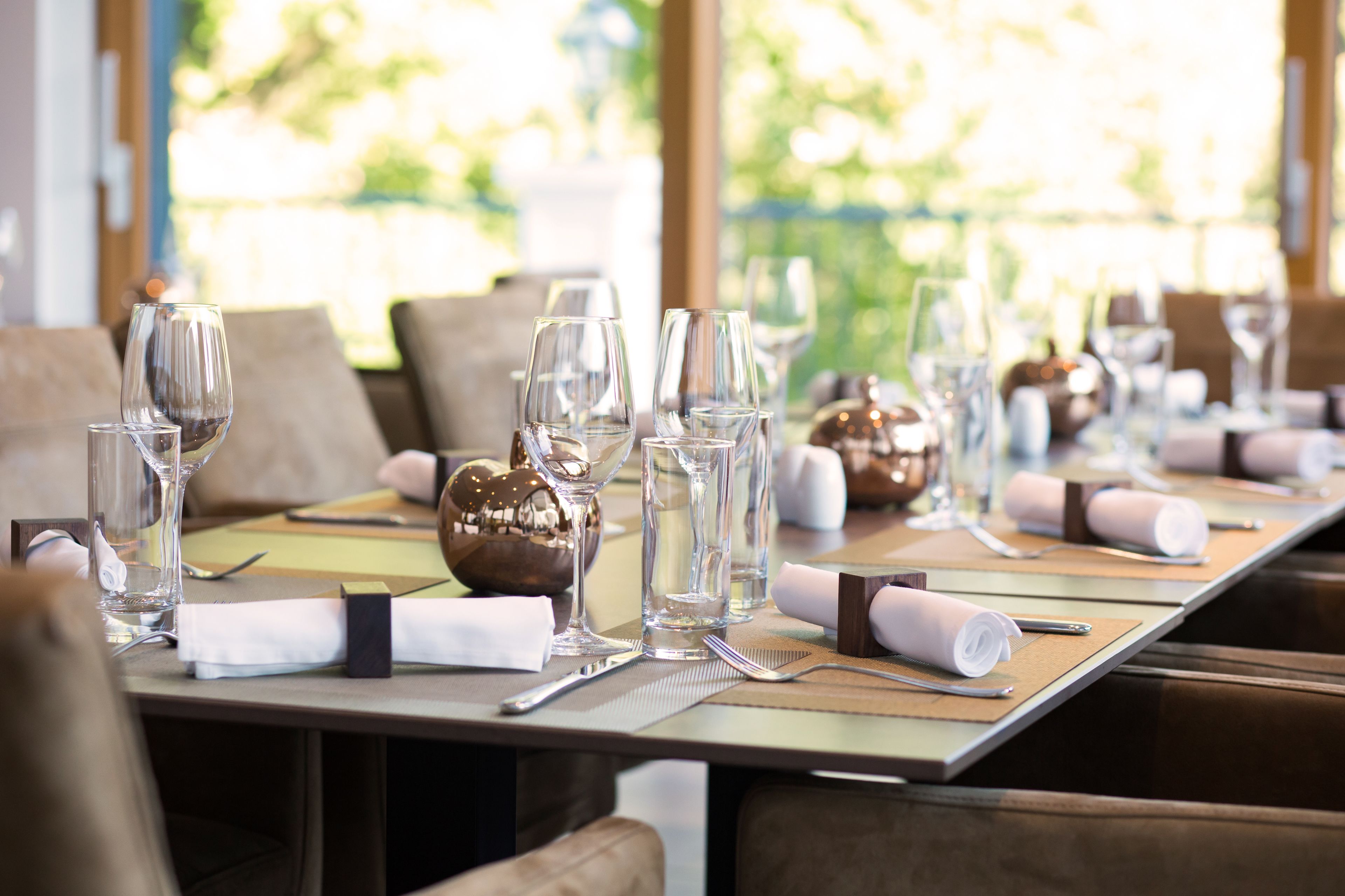 A dining table set with multiple wine glasses, water glasses, and cutlery, with a view of greenery outside.