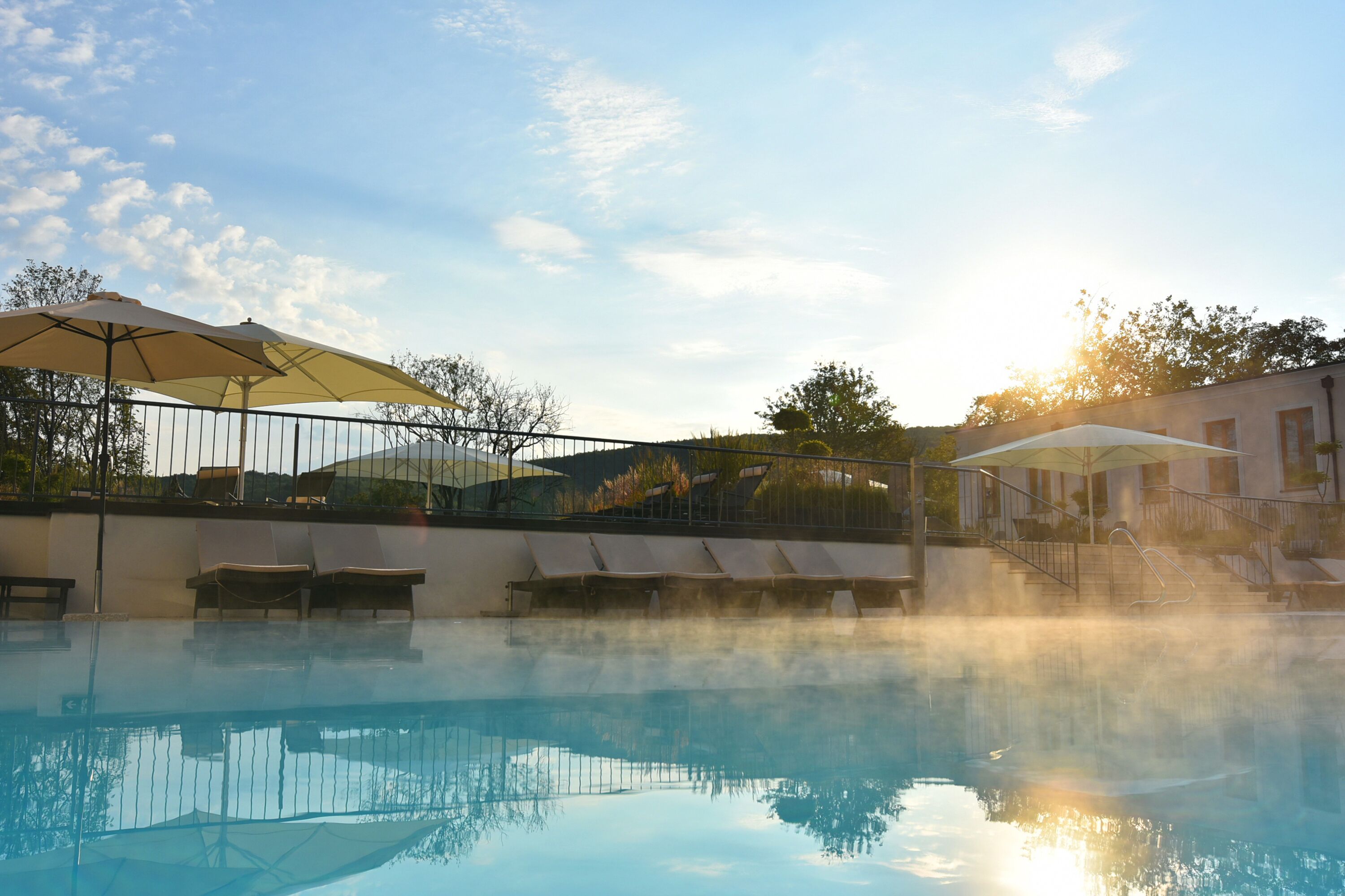 A swimming pool with lounge chairs and umbrellas, surrounded by a fence and trees.