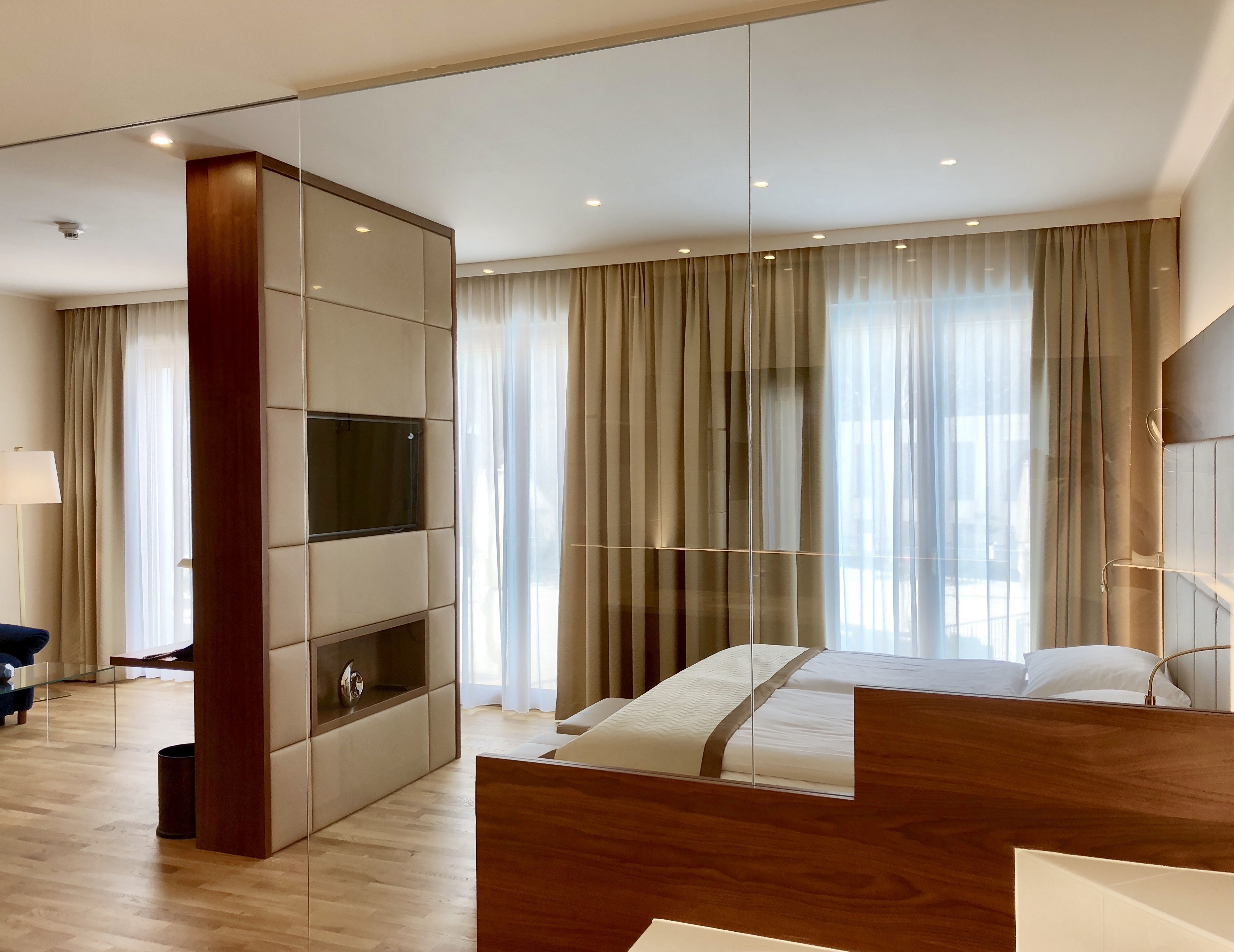 Ein modernes Hotelzimmer mit einem großen Bett, einem an einer Holzeinrichtung montierten Fernseher und Blick auf die Stadt durch große Fenster mit leichtem Vorhang.
