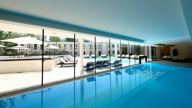 Une piscine intérieure moderne avec des parois en verre transparent, des chaises longues et des parasols.