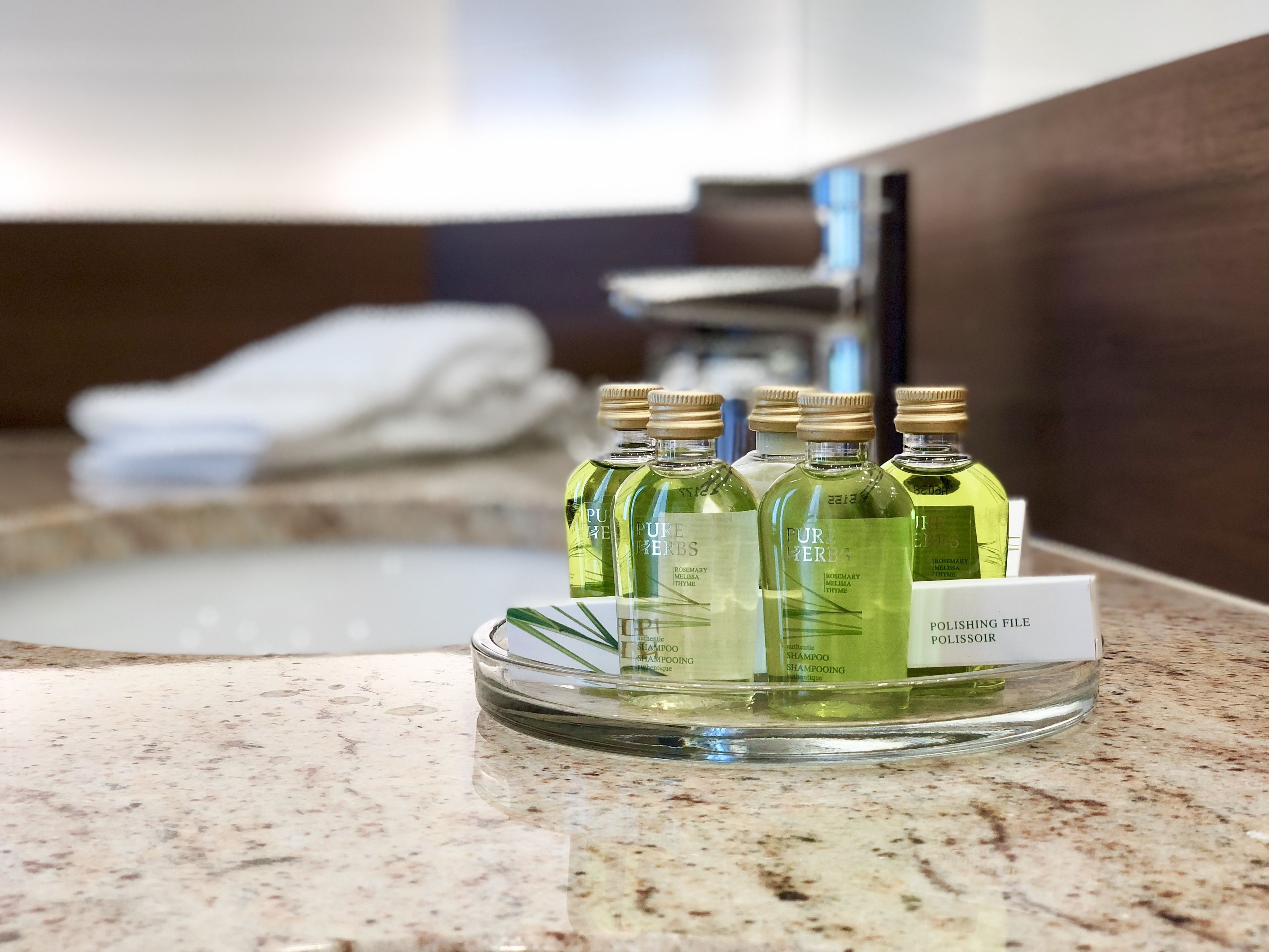 A bathroom with a granite countertop, a glass dish holding several bottles of Pure Herbs shampoo, and a white towel hanging in the background.