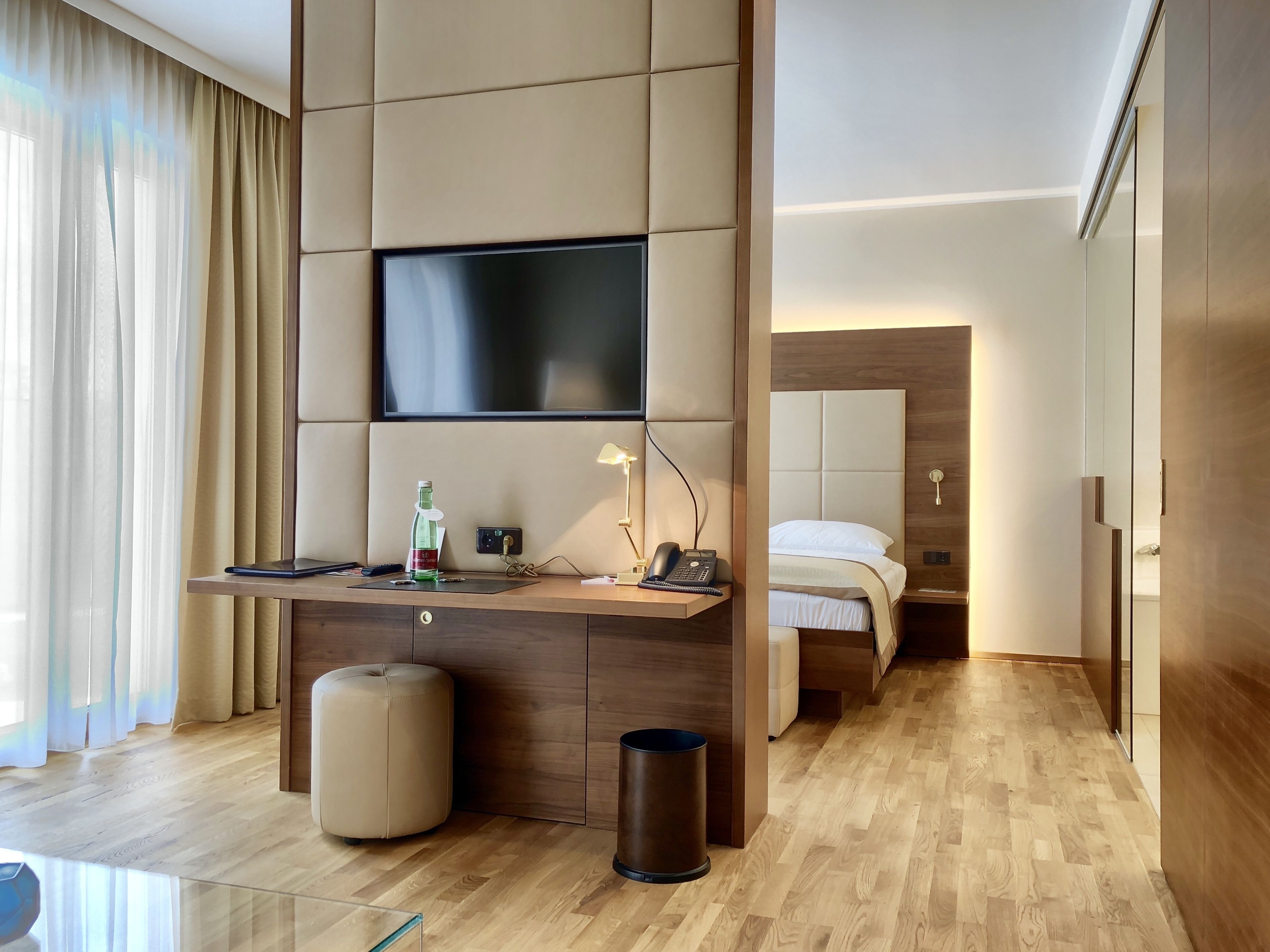 Een moderne hotelkamer met een houten vloer, een grote flatscreen televisie aan de muur, een bureau met een lamp, een nachtkastje en een bed met hoofdeinde.