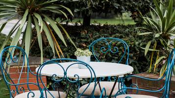 A garden setting with a white table and blue metal chairs, surrounded by lush greenery and a large bush.