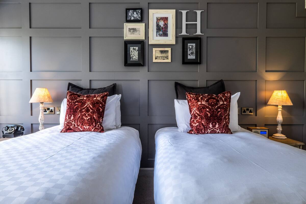 A hotel room with two beds, each with a patterned pillow and a white bedspread. There are framed pictures on the wall above the beds and a large 'H' above them.