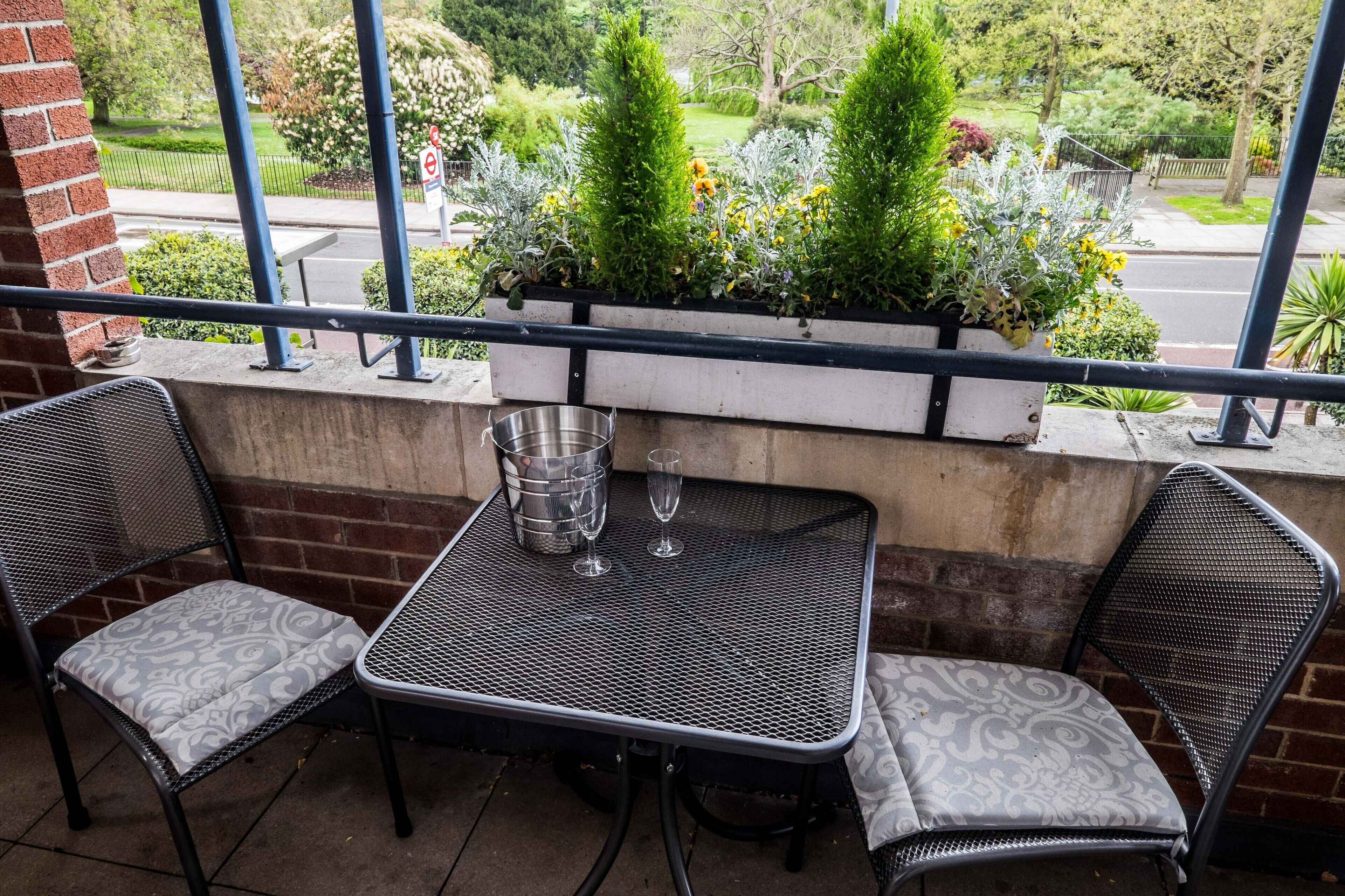 A balcony with two metal chairs and a table, a metal bucket, and a potted plant with flowers.