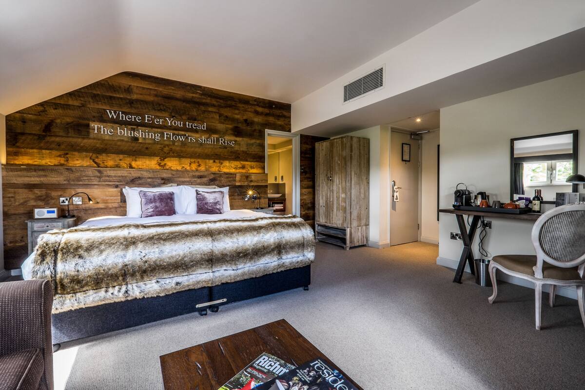A bedroom with a large bed, a wooden accent wall, a desk with a chair, and a small table with magazines.