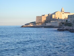 A coastal town with historic buildings, including a prominent church with a bell tower, situated on a rocky shoreline. - Residence Fontana Vecchia (Vieste)