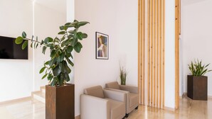 Un hall d’entrée moderne agrémenté d’une grande plante en pot, de deux canapés beiges, d’une cloison en lattes de bois et d’un tableau abstrait encadré au mur.