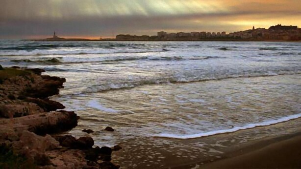 Un paysage côtier avec des vagues qui s’écrasent sur le rivage, une côte rocheuse et, au loin, le profil d’une ville sous un ciel nuageux.