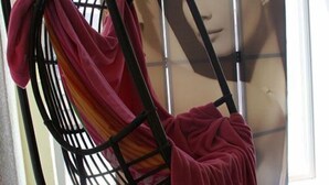Een kamer met een ronde tafel bedekt met een rode tafelkleed, een hangstoel met een roze deken, en een poster aan de muur.