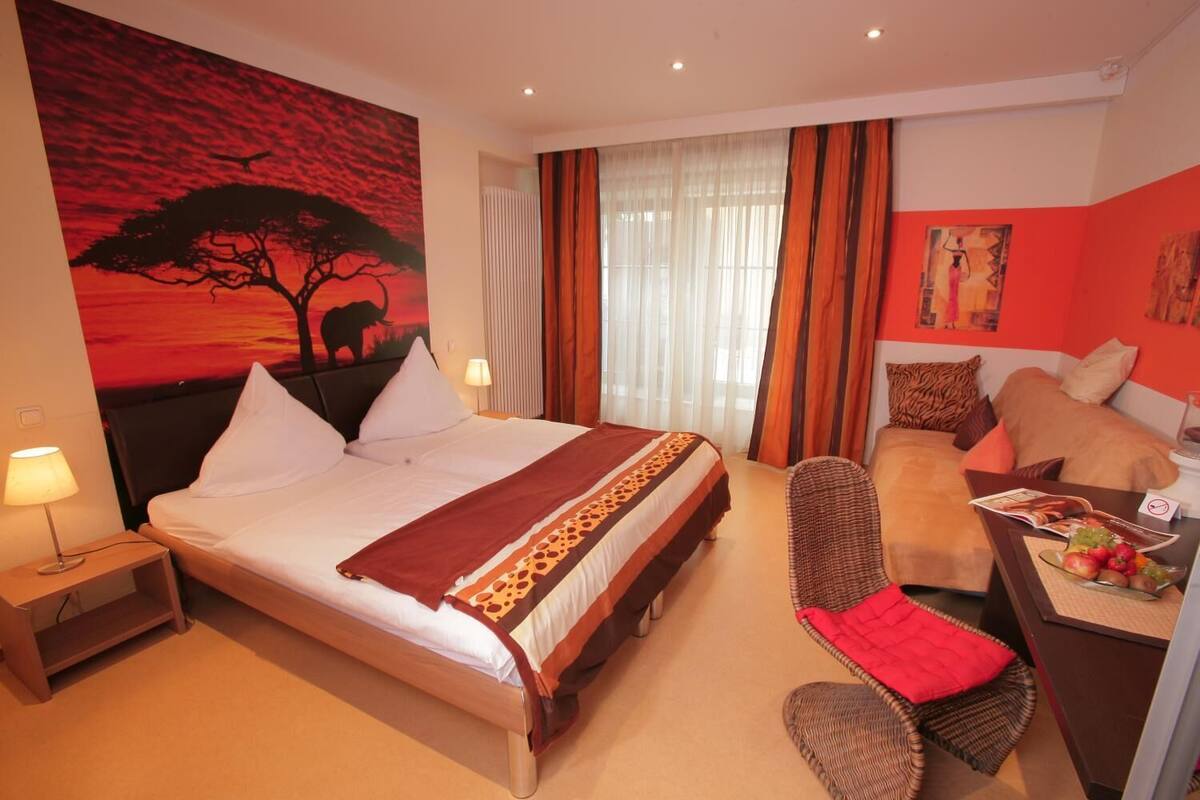 A hotel room with a large bed, a desk with a chair, a dining table with fruit, and a painting of a tree.