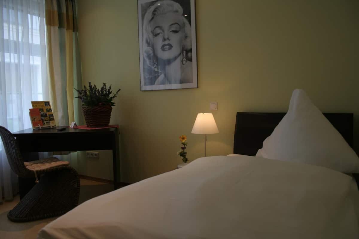 A hotel room with a bed, a desk with a lamp, a chair, a plant, and a framed picture on the wall.
