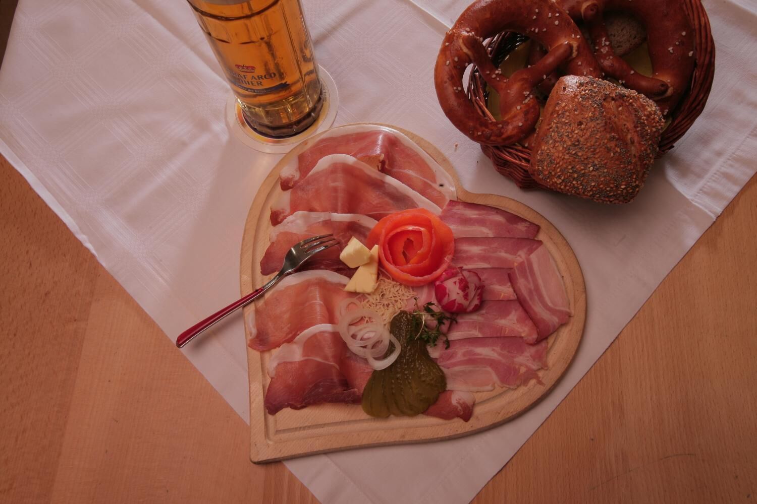 Una bandeja con forma de corazón, con diversas carnes, un pretzel y una jarra de cerveza sobre una mesa.