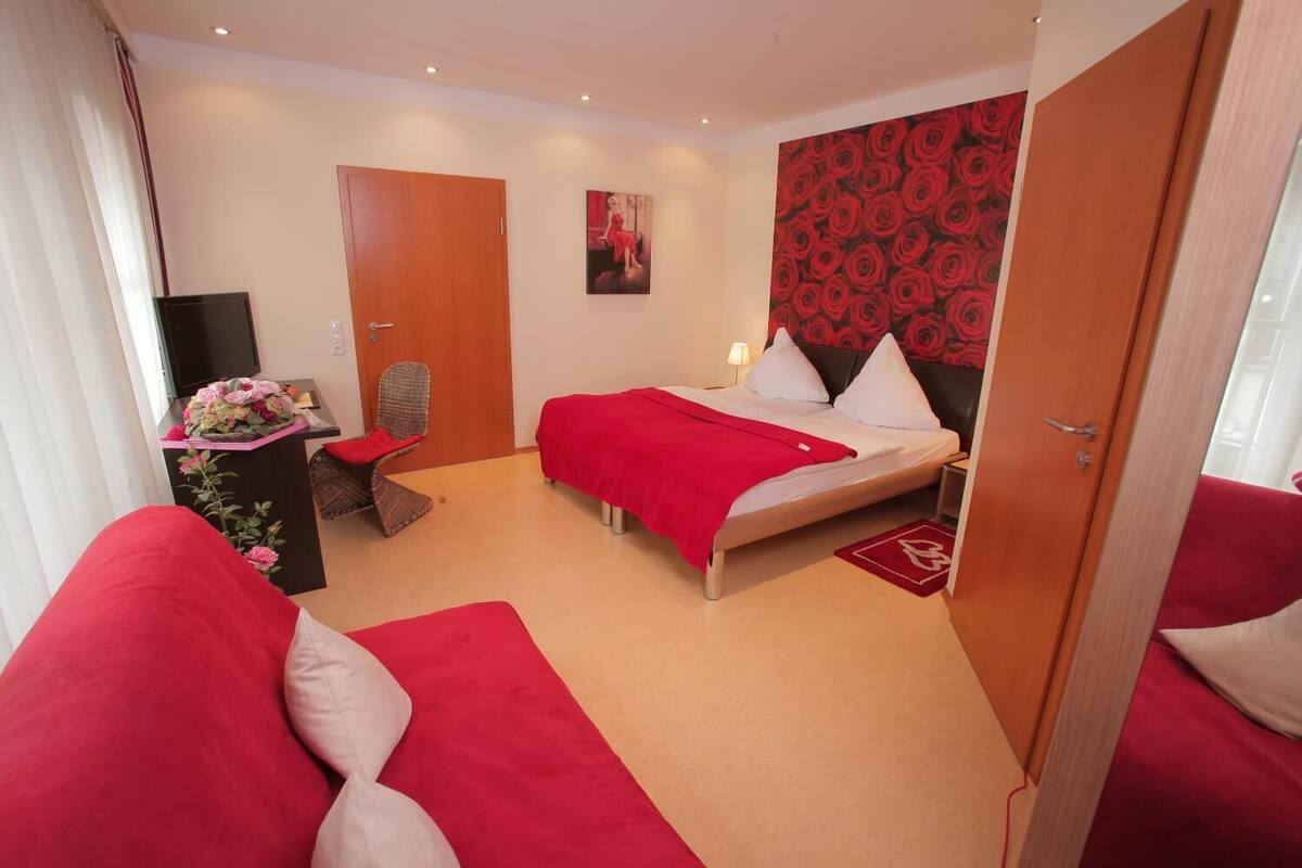 A hotel room with a bed, a chair, a desk with a TV, and a red floral arrangement.