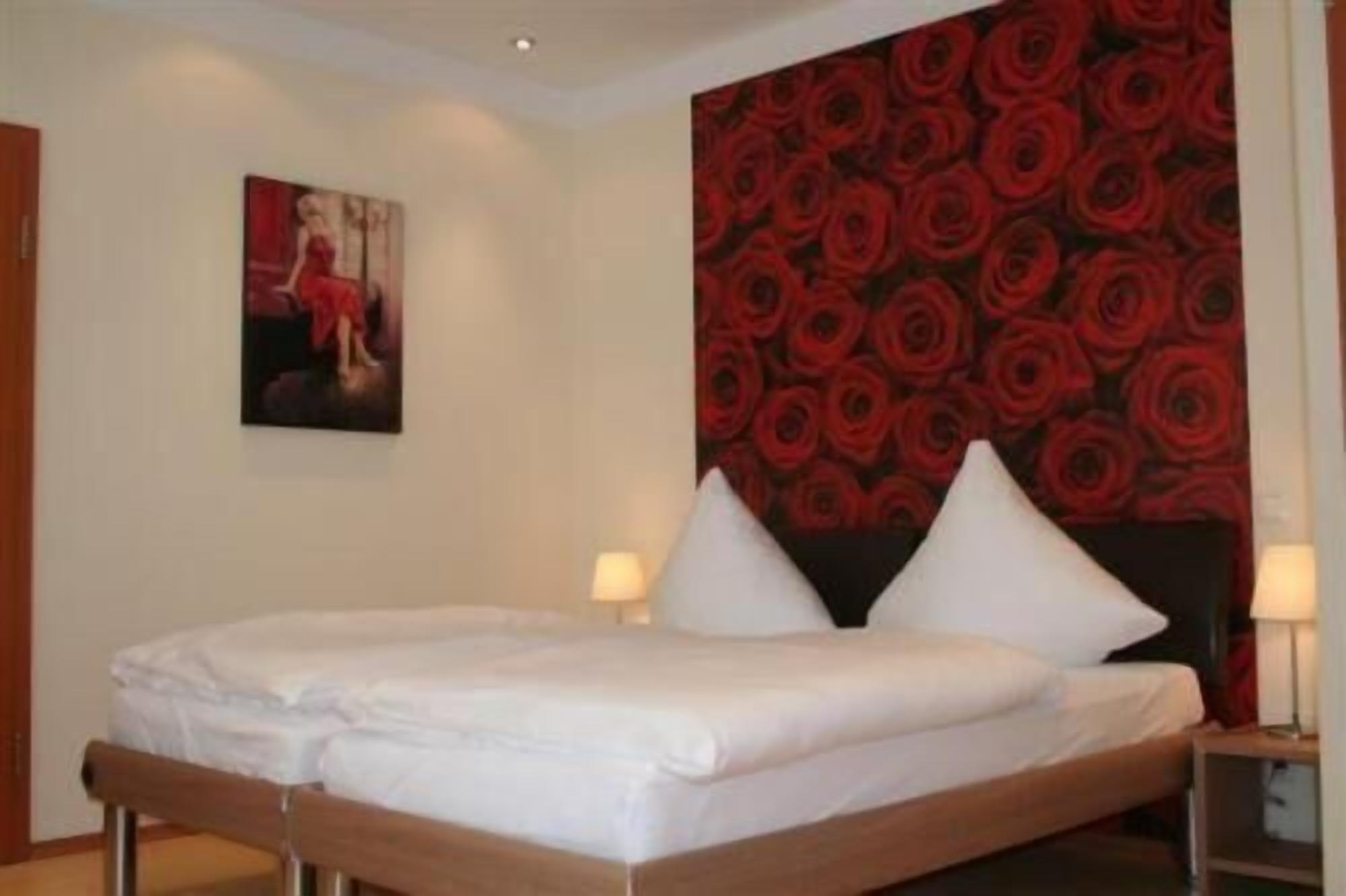 Una cama doble con sábanas blancas, dos mesitas de noche con lámparas y un papel tapiz floral rojo en la pared.