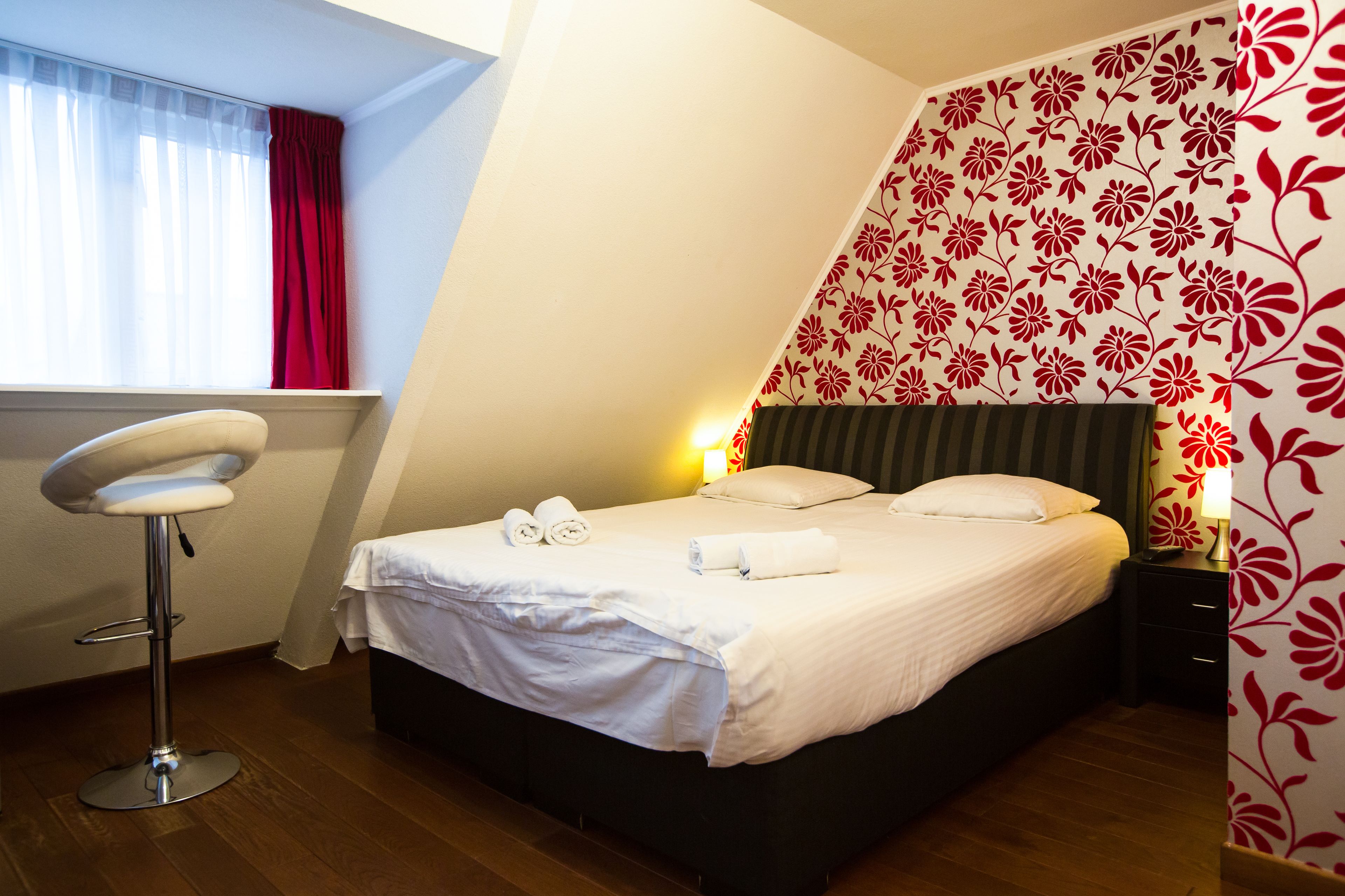 Une chambre à coucher avec un lit, une table de chevet, une lampe, une fenêtre avec des rideaux et une chaise.