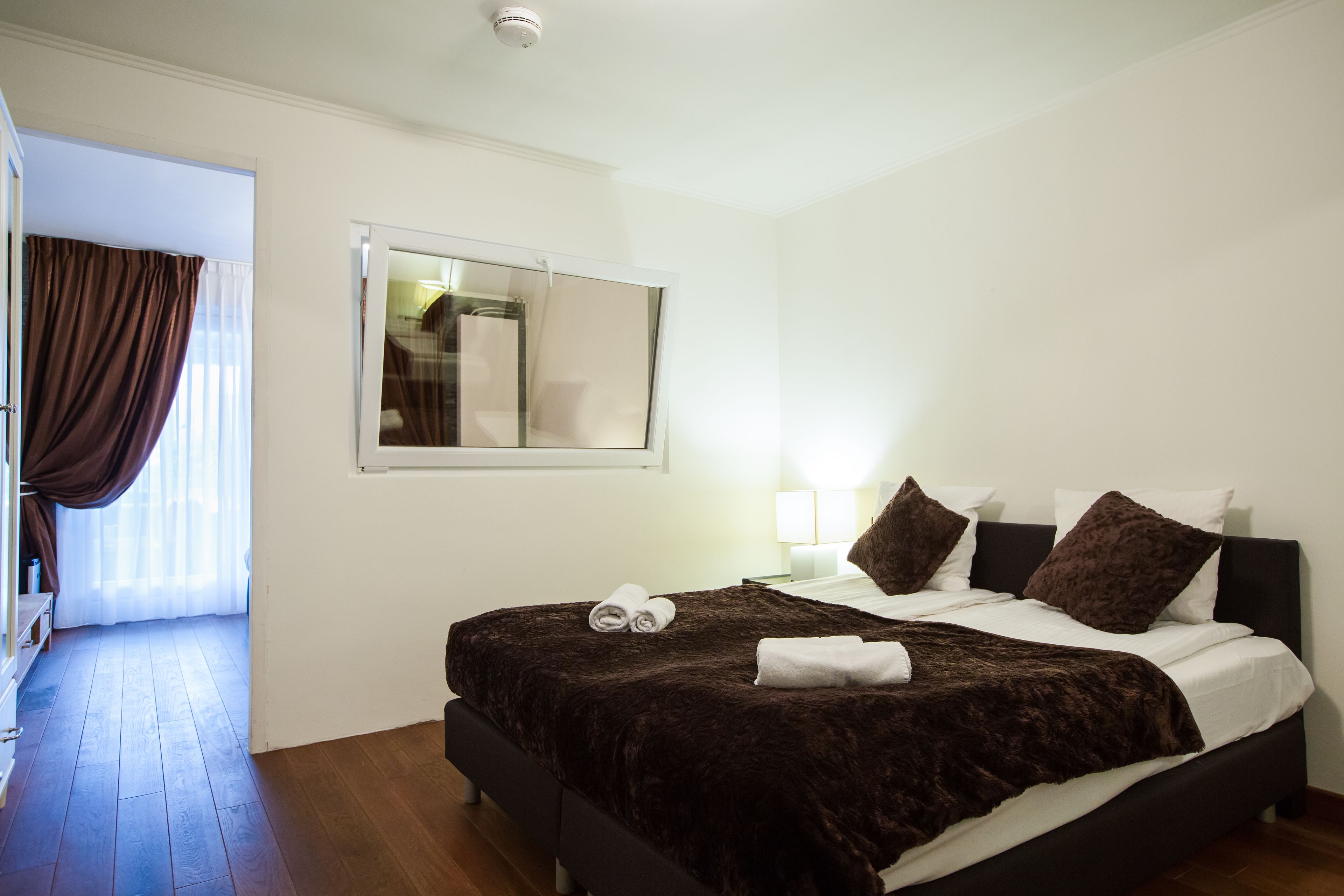 Un dormitorio con una cama grande, una ventana con cortinas, una mesita de noche con lámpara y un espejo en la pared.