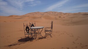 A desert landscape with sand dunes in the background and a metal table with chairs in the foreground. - Riad Aicha (Taouz)