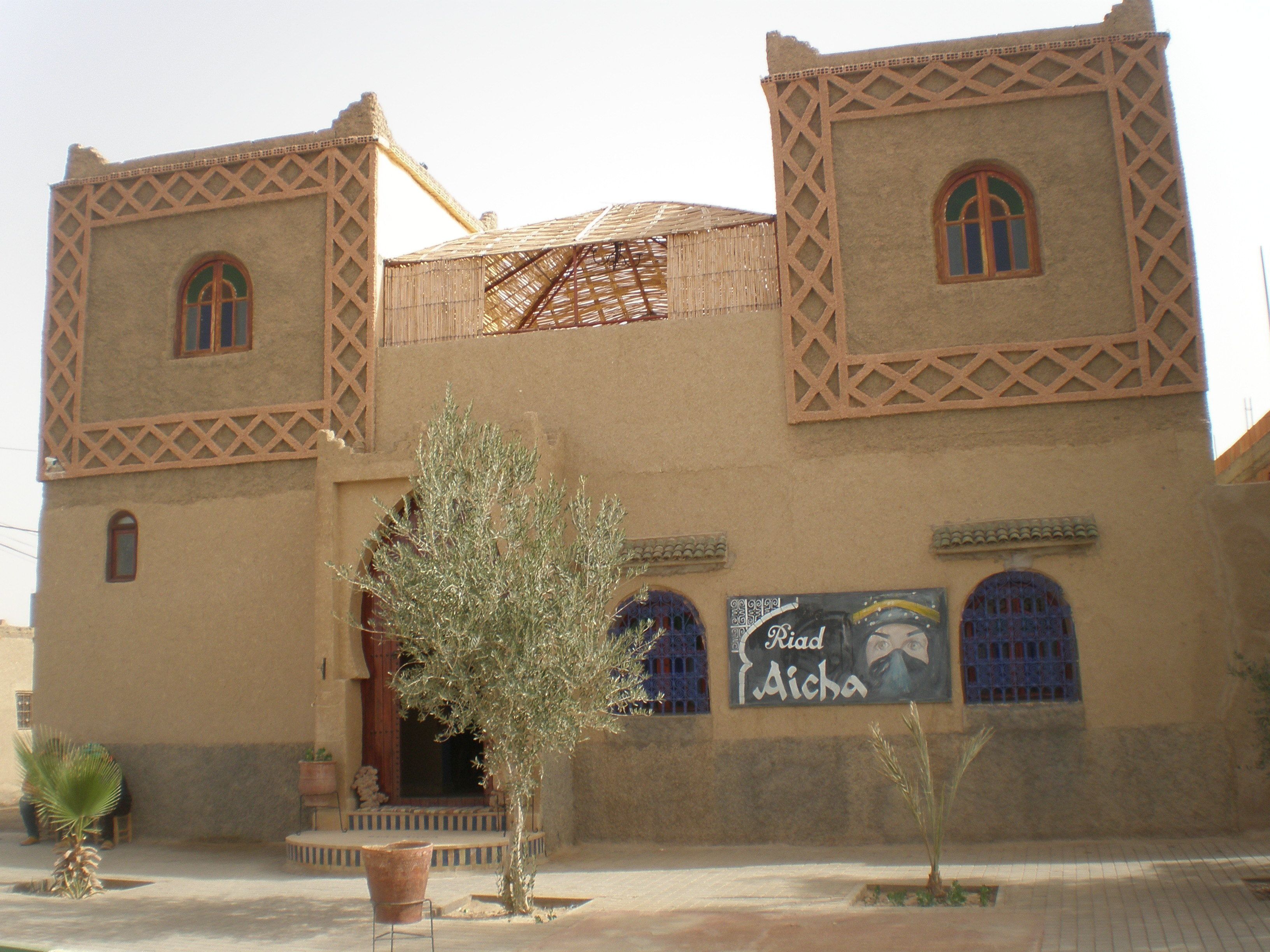 A traditional building with two towers, a central entrance, and a sign reading "Riad Aicha."