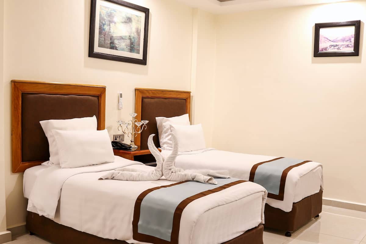 Quarto de hotel com duas camas, cada uma com cabeceira de madeira, roupa de cama branca e detalhes em castanho. Há quadros nas paredes e uma mesa de cabeceira com abajur e telefone.