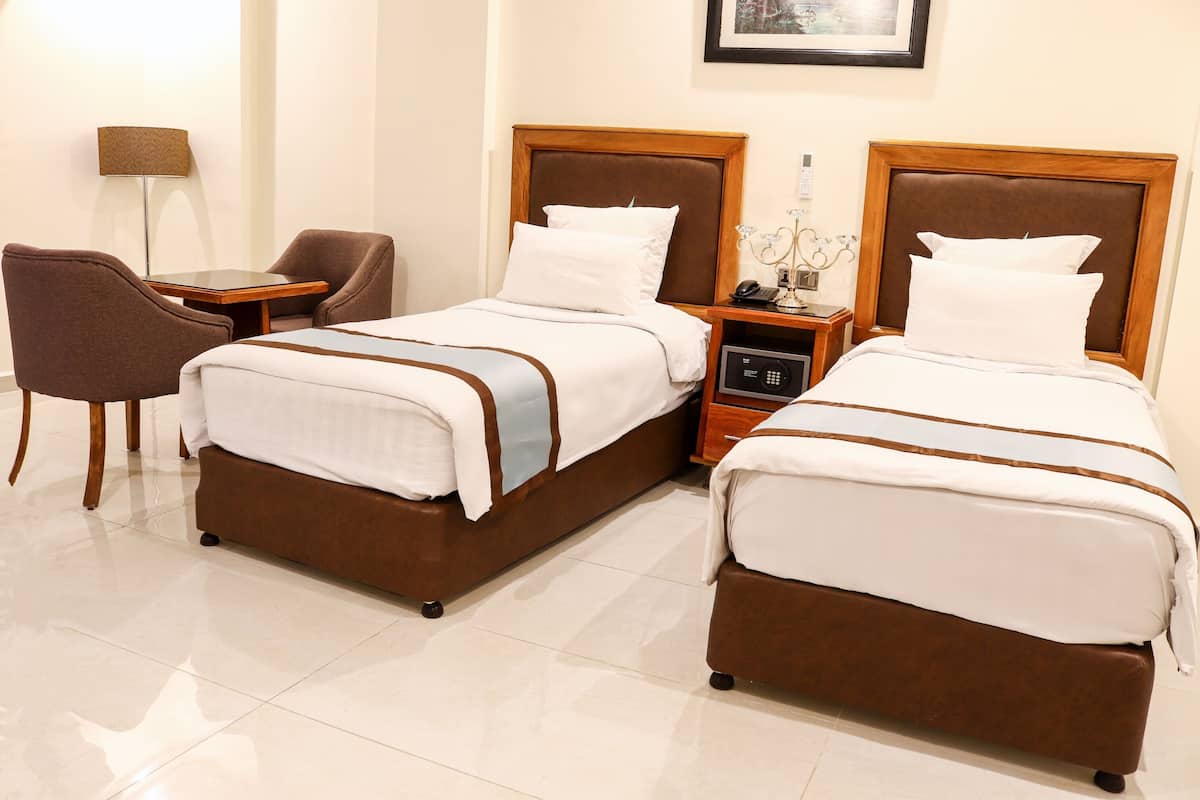 Quarto de hotel com duas camas, uma escrivaninha, uma cadeira, uma mesa de cabeceira com abajur e um quadro na parede.