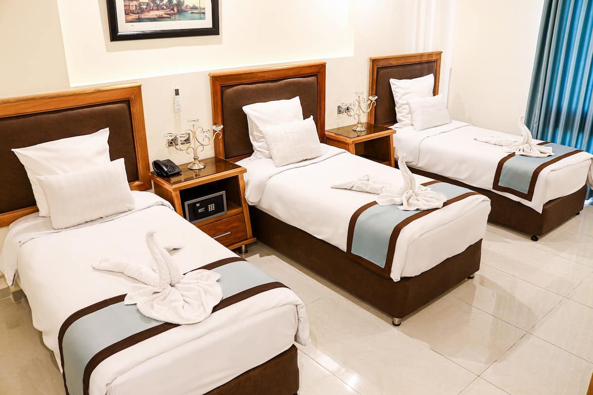 Quarto de hotel com três camas, cada uma com cabeceira de madeira, roupa de cama branca e uma toalha sobre a cama. Há uma mesa de cabeceira com telefone e abajur entre as camas.