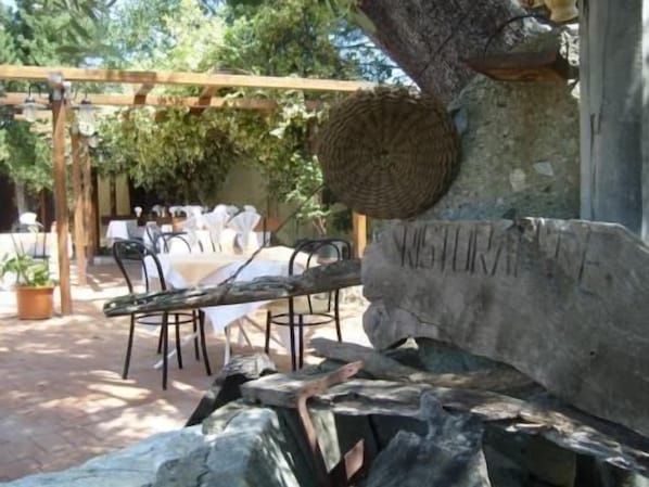 Un área de comedor al aire libre con una rústica pared de piedra, pérgola de madera y mesas dispuestas para comer.