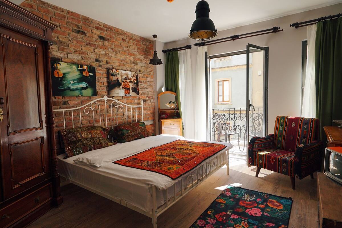 A bedroom with a brick wall, a bed with colorful bedding, a vintage TV, and a balcony with a chair.