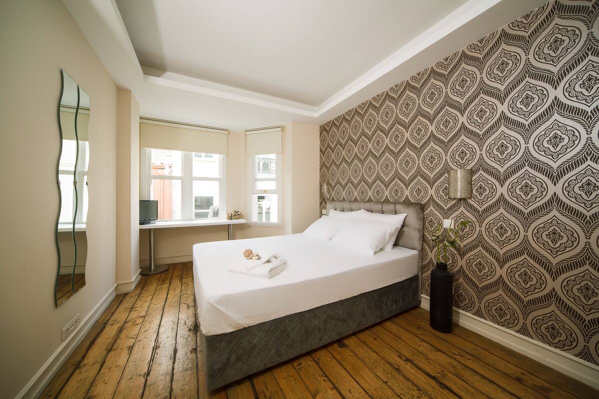 A bedroom with a bed, a desk, a mirror, and patterned wallpaper.