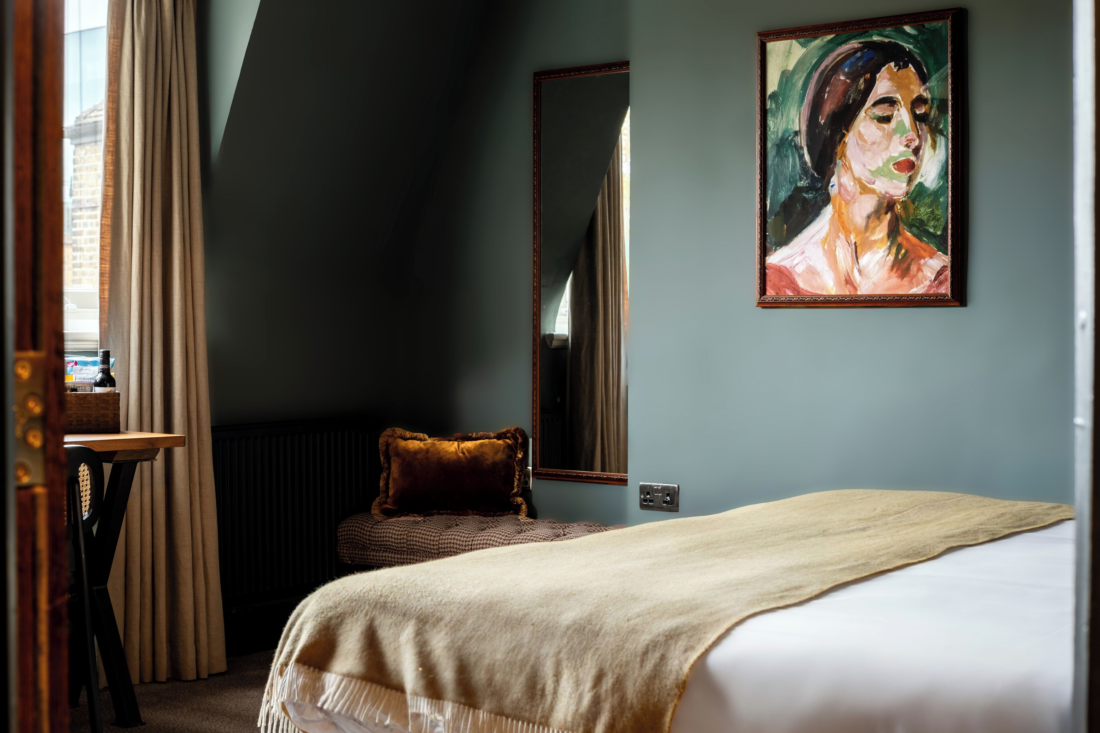 Ein Hotelzimmer mit Bett, einem Gemälde an der Wand, einem Fenster mit Vorhängen und einem kleinen Tisch mit Lampe.