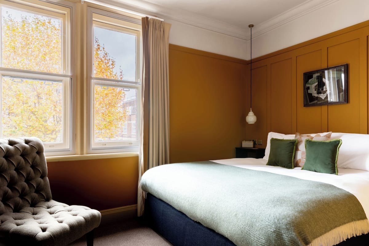 A hotel room with a large bed, a tufted armchair, a nightstand, and a window with curtains.