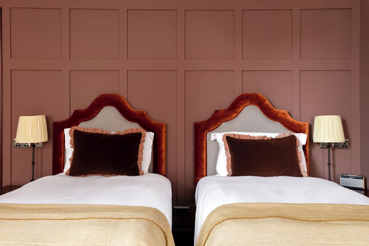 Two identical twin beds with a plush headboard, white bedding, and a beige throw blanket. Each bed has a bedside lamp and a small table with a radio.