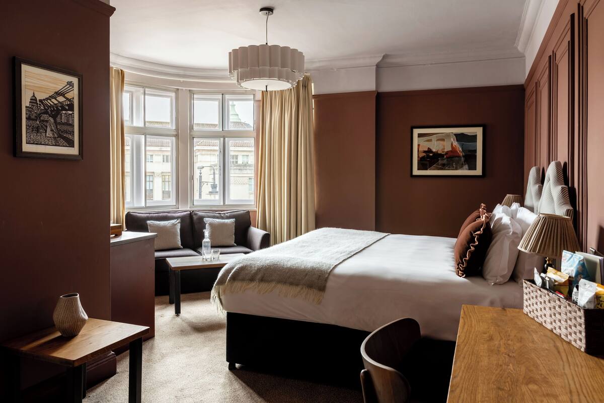 A hotel room with a large bed, a desk, a sofa, a window with curtains, and artwork on the walls.