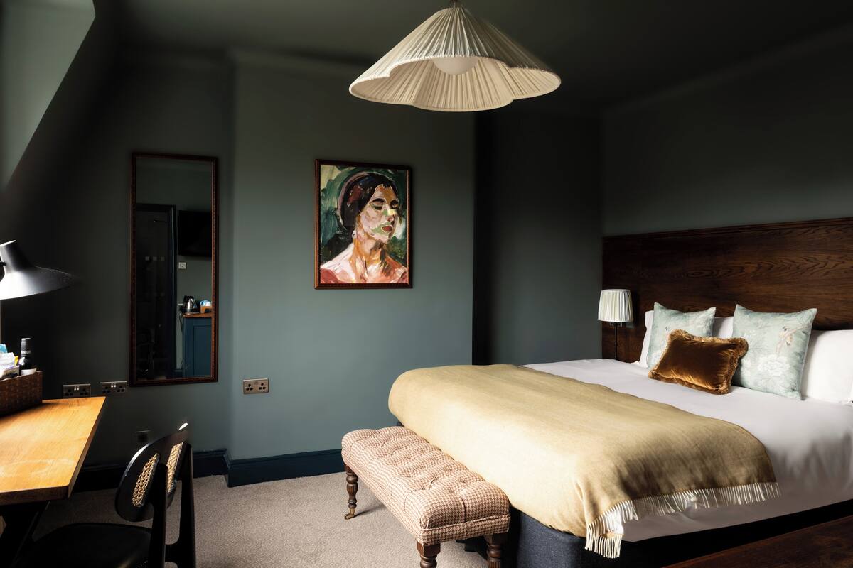A bedroom with a large bed, a wooden headboard, a bedside table, a lamp, a bench, and a painting on the wall.