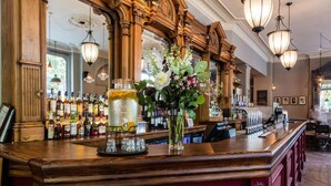 A bar area with a wooden counter, various bottles, a large jar, and a vase with flowers. - Brook Green Hotel (London)