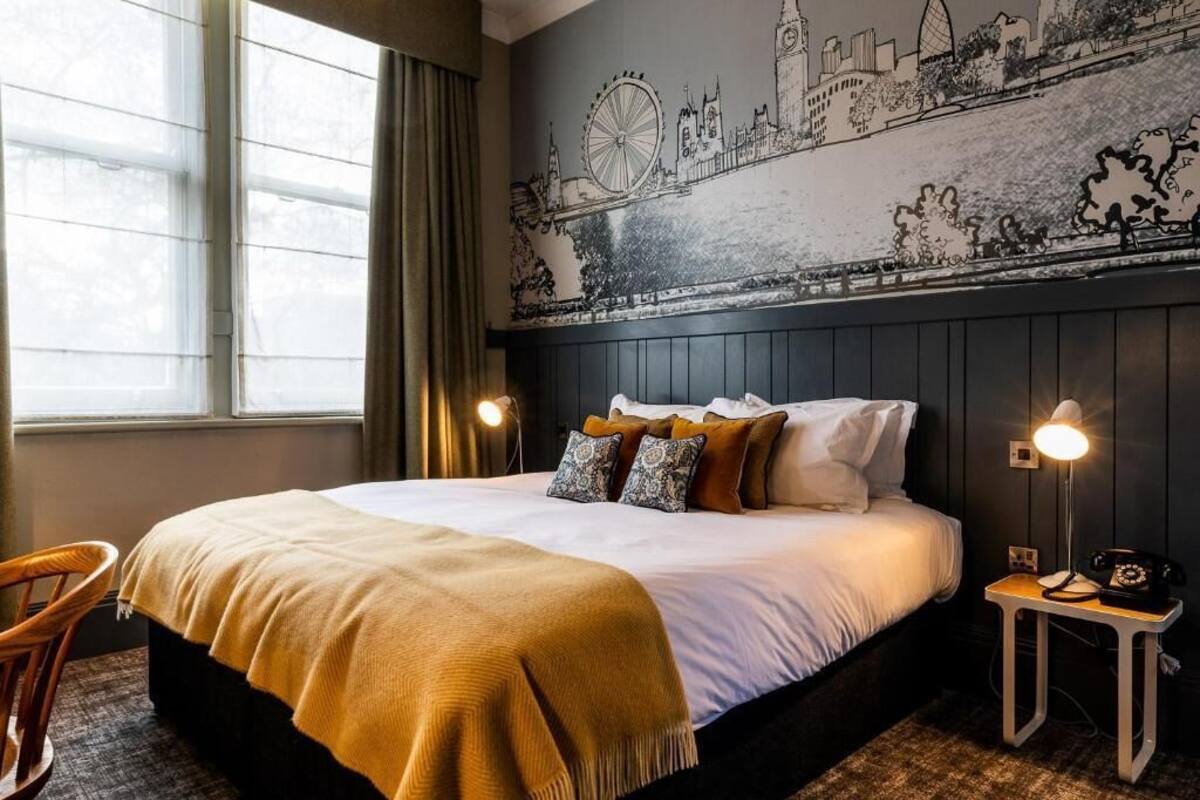 Hotellihuone, jossa on suuri sänky, kaksi yöpöytävalaisinta, tuoli ja seinällä kaupunkimaisemaa esittävä seinämaalaus.