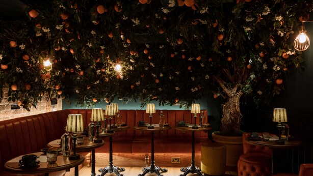 A dimly lit lounge area with round tables, leather seating, and a large artificial tree with hanging oranges. - Brook Green Hotel (London)
