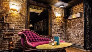 A room with a brick wall, a plush pink sofa, a wooden table, and a mirror. - Brook Green Hotel (London)