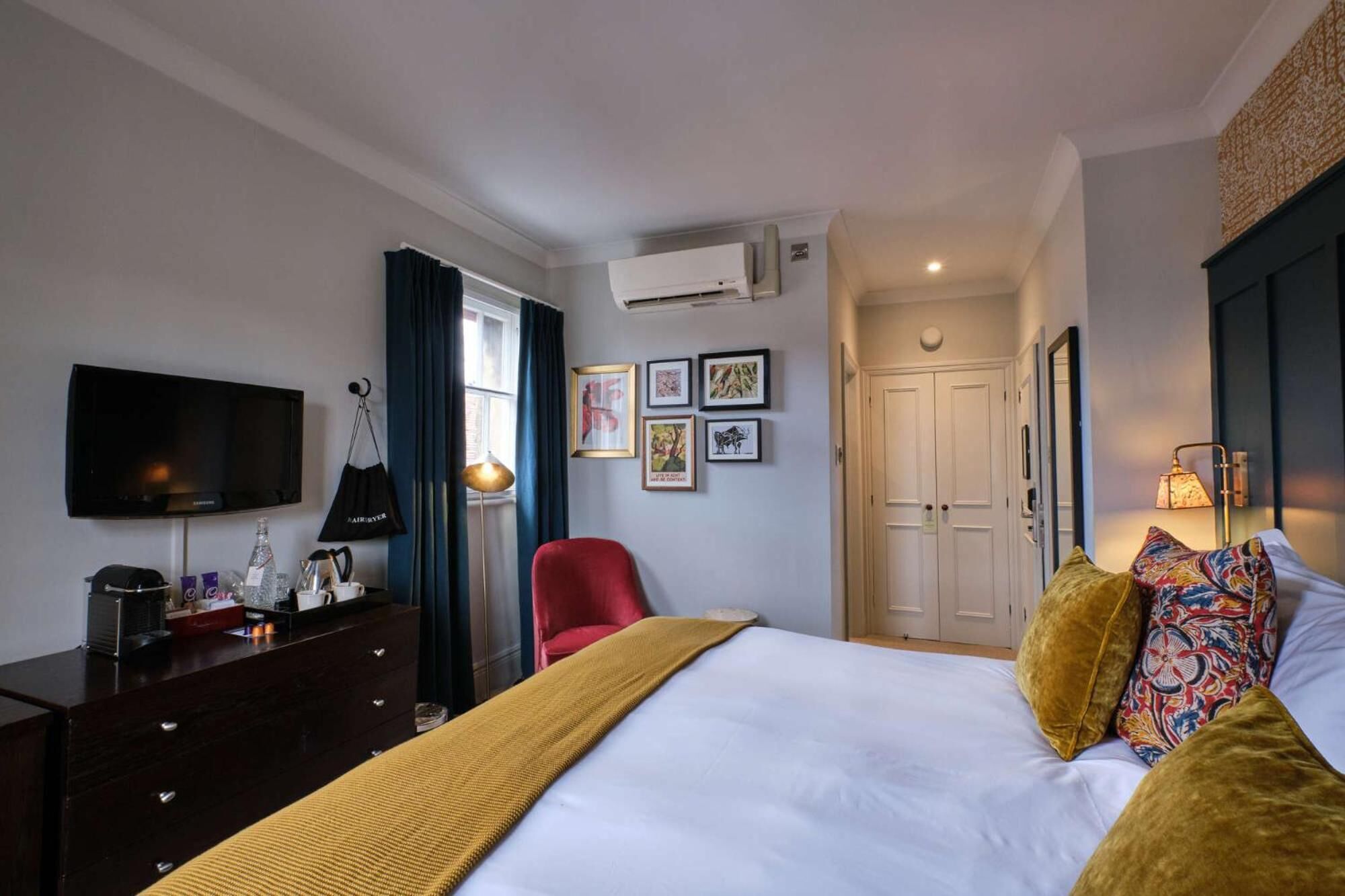 A hotel room with a bed, a TV, a dresser, a red chair, a desk with a lamp, and a door leading to another room.