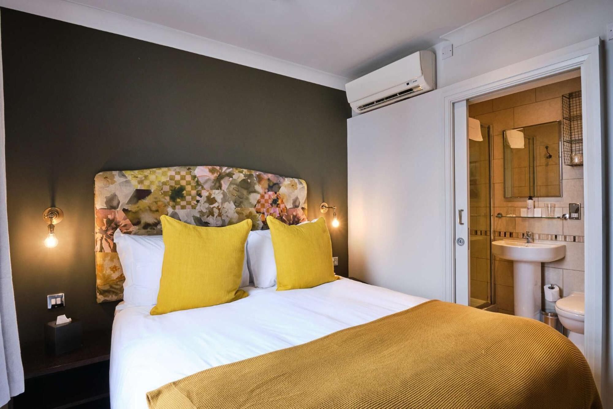 A hotel room with a bed, two yellow pillows, a floral headboard, a white blanket, an air conditioning unit, and a bathroom with a sink and mirror.