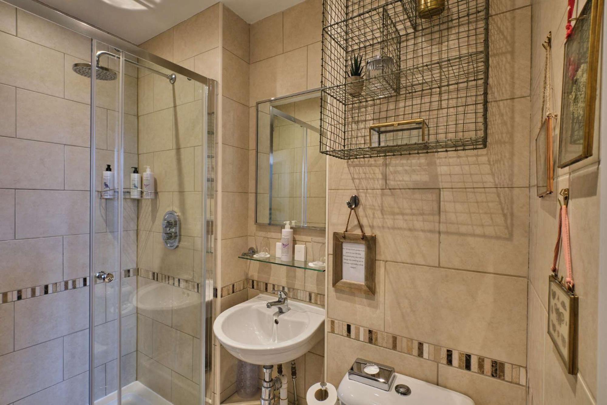A bathroom with a glass-enclosed shower, a white sink, and a toilet. There are toiletries on the sink and a framed note on the wall.