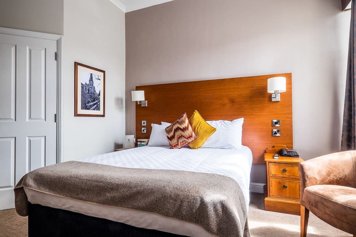 A hotel room with a large bed, a wooden headboard, two bedside tables with lamps, a chair, and a framed picture on the wall.