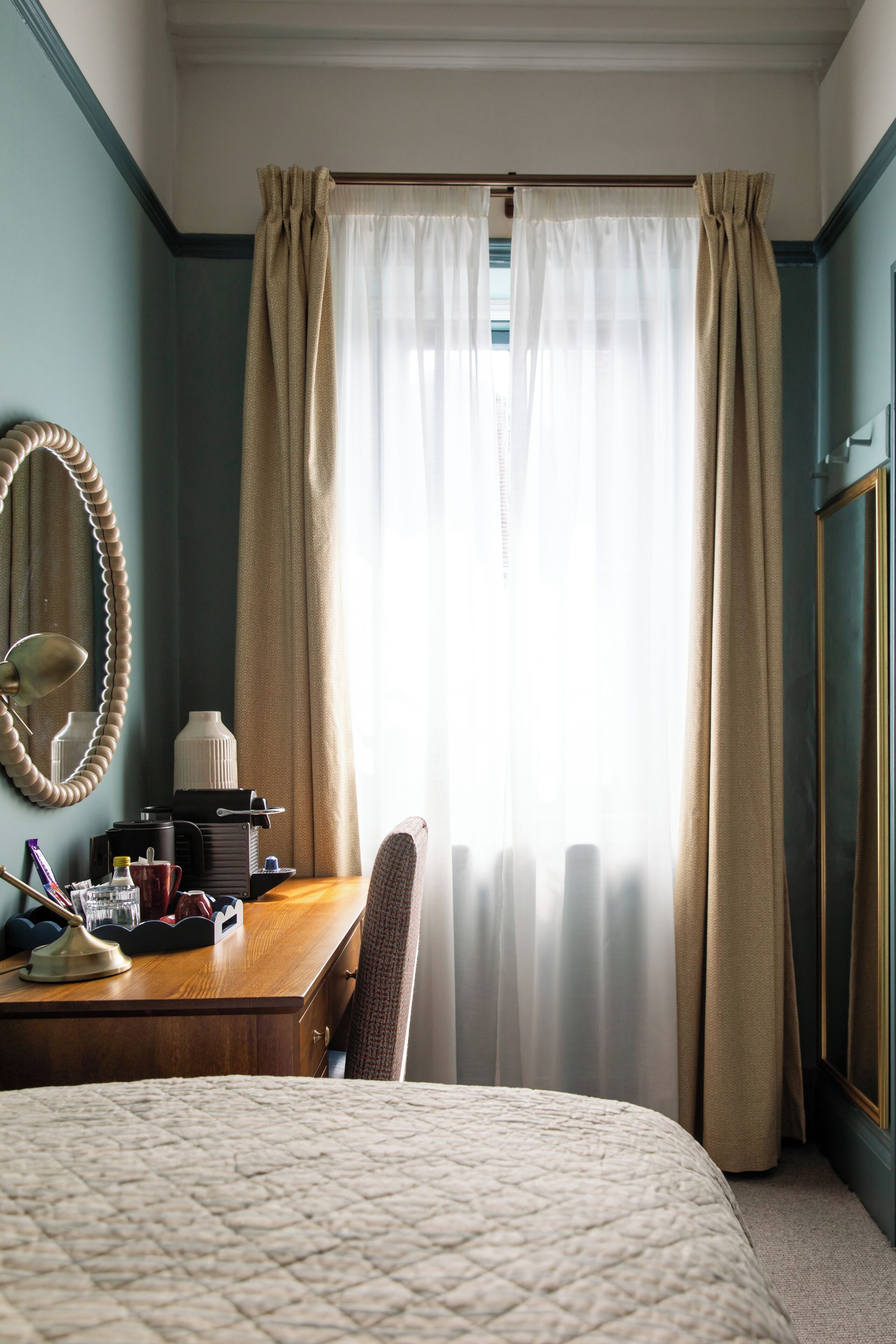 A hotel room with a bed, a desk with a lamp, a chair, a mirror, and a window with curtains.