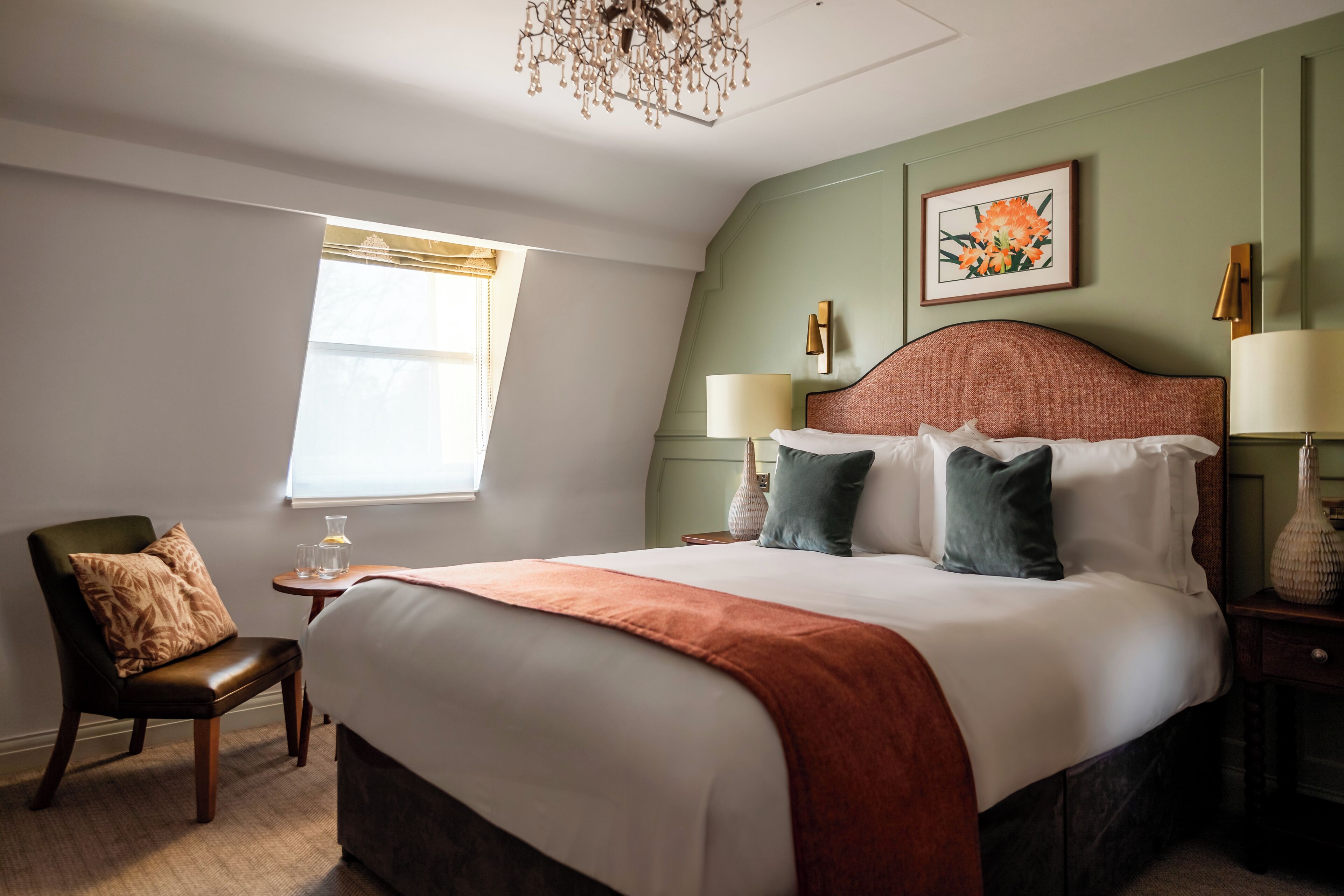 A neatly arranged bedroom with a large bed, a chair, a bedside table with a lamp, and a window with curtains.