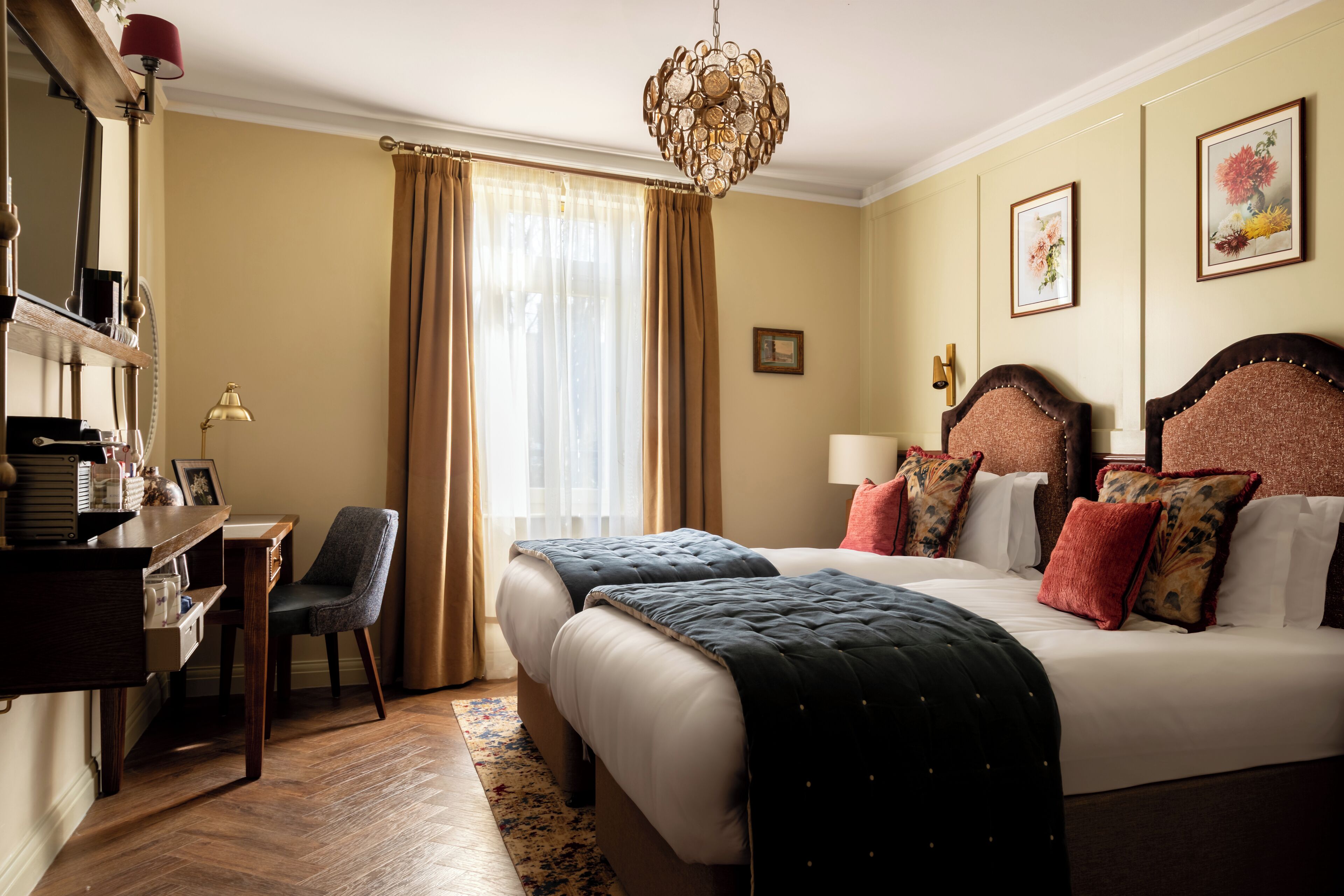 A hotel room with two beds, a desk, a chair, a desk lamp, a chandelier, and framed artwork on the walls.