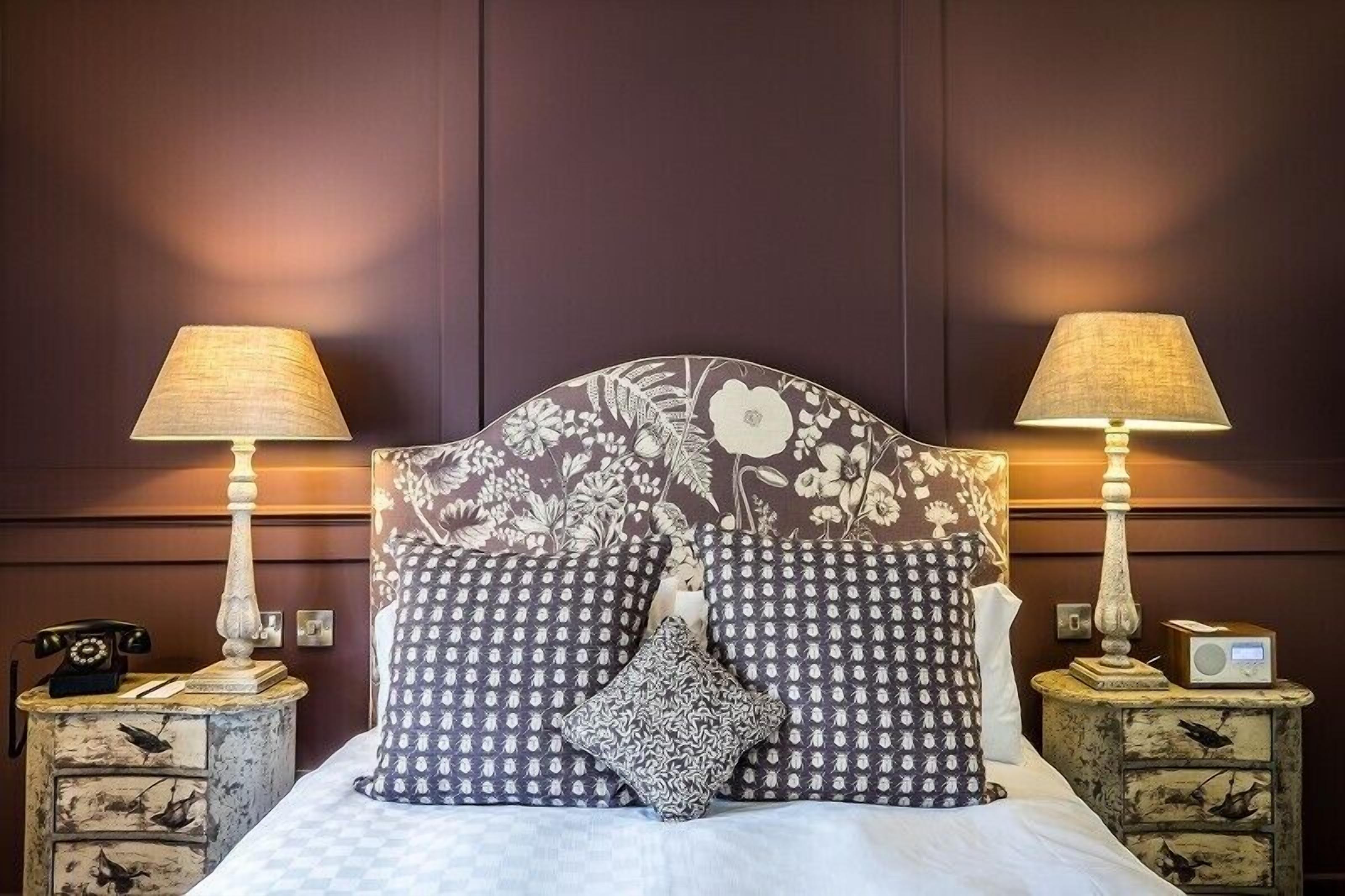 A bed with a floral headboard, two bedside tables with lamps, a telephone, and a radio on one of the tables.