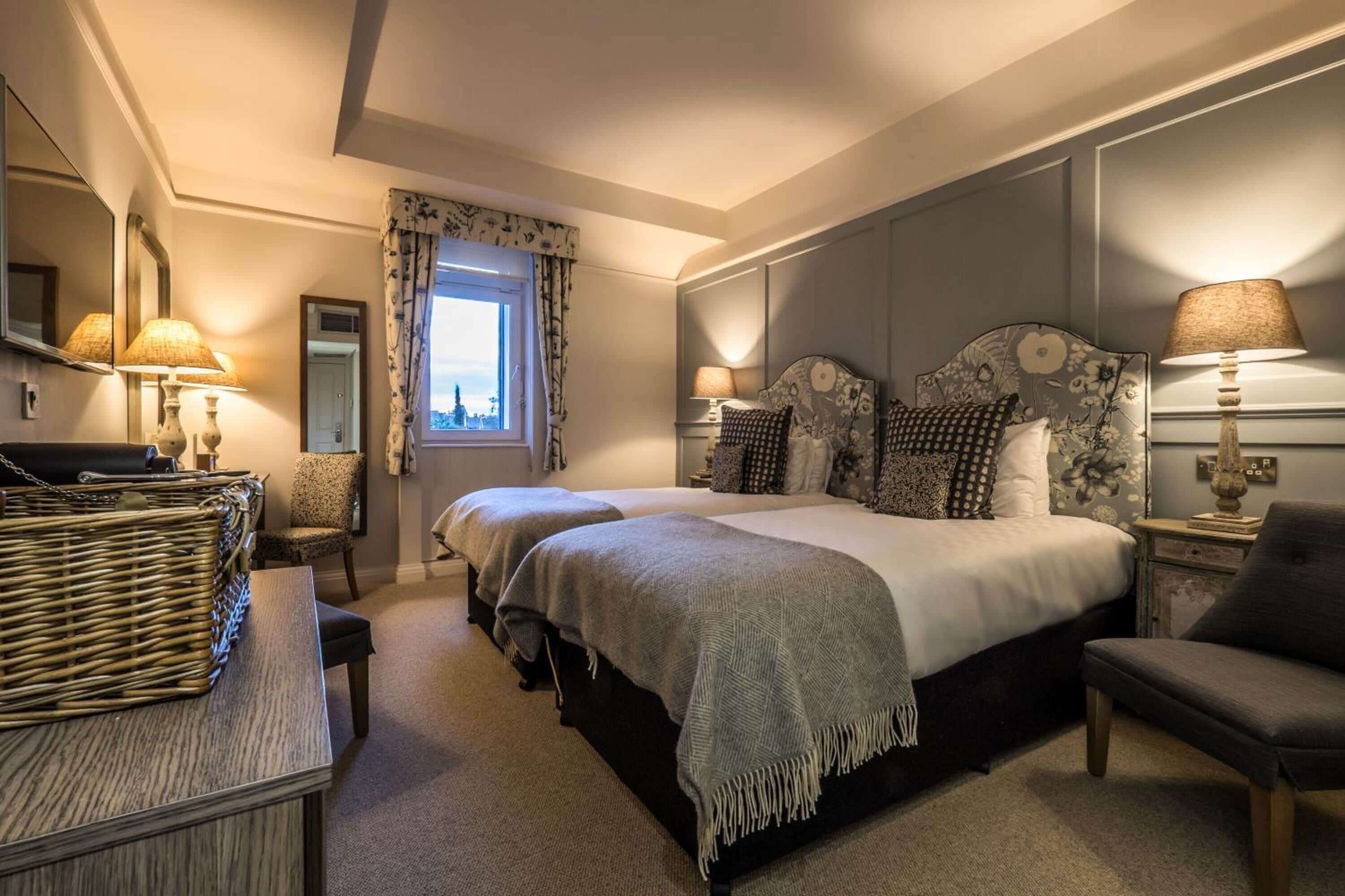Quarto de hotel com duas camas, uma janela grande com cortinas florais, uma mesinha de vime e uma cadeira.