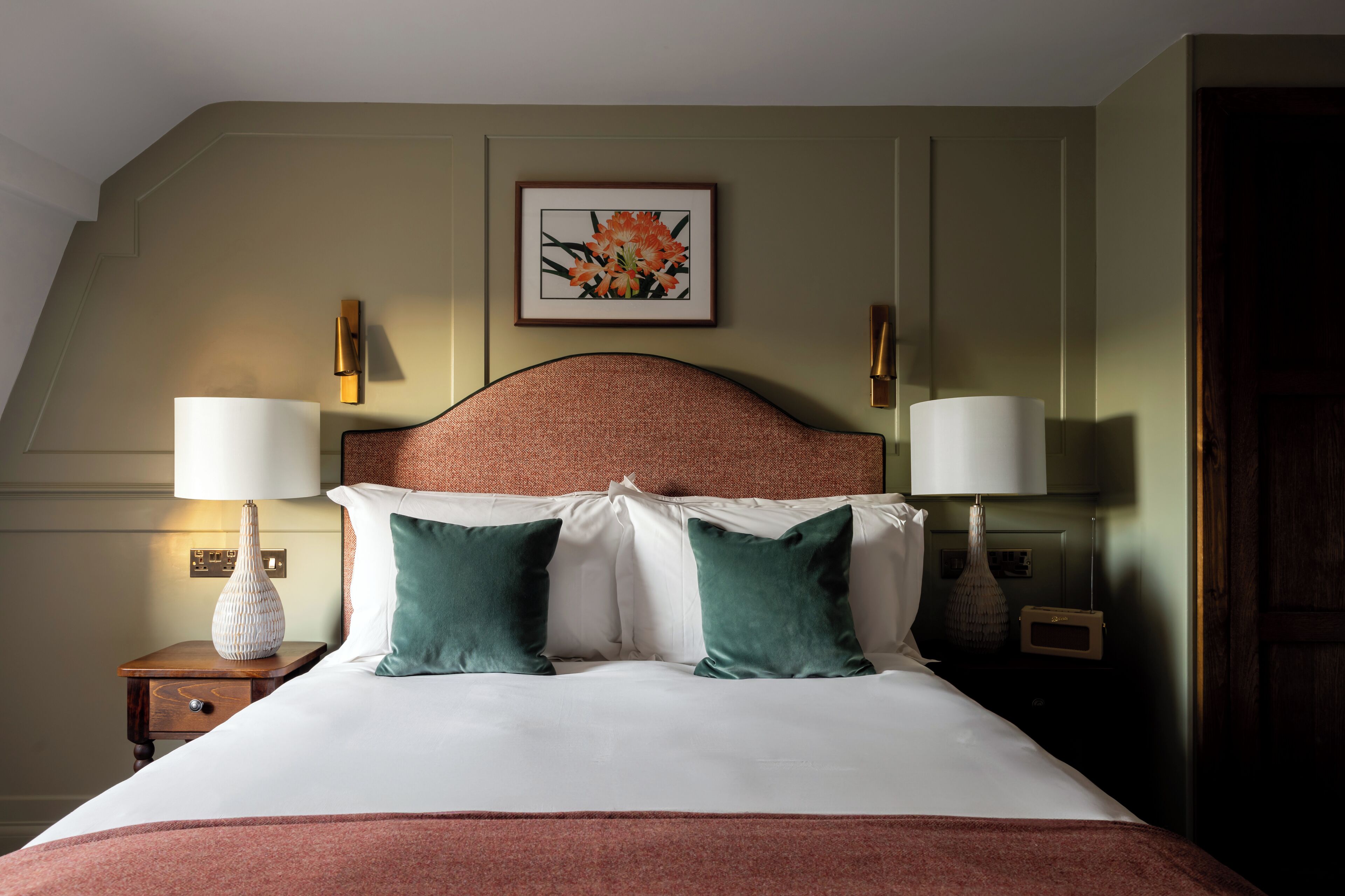 A neatly made bed with a headboard, two bedside lamps, and a framed floral artwork on the wall.