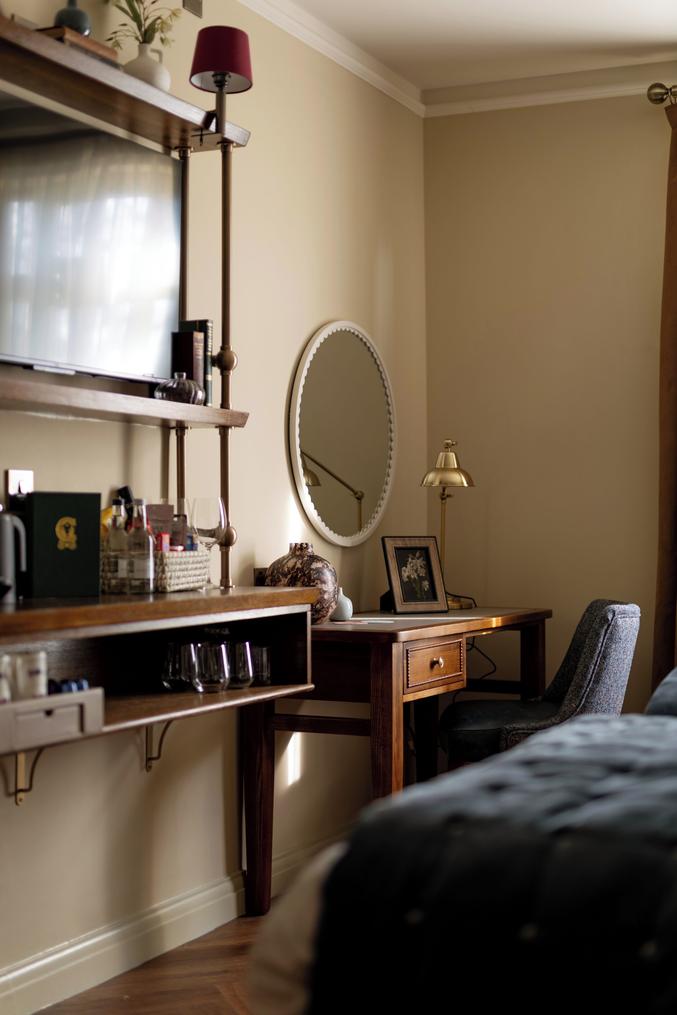 A bedroom with a bed, a desk with a lamp, a mirror, and a shelf with various items.