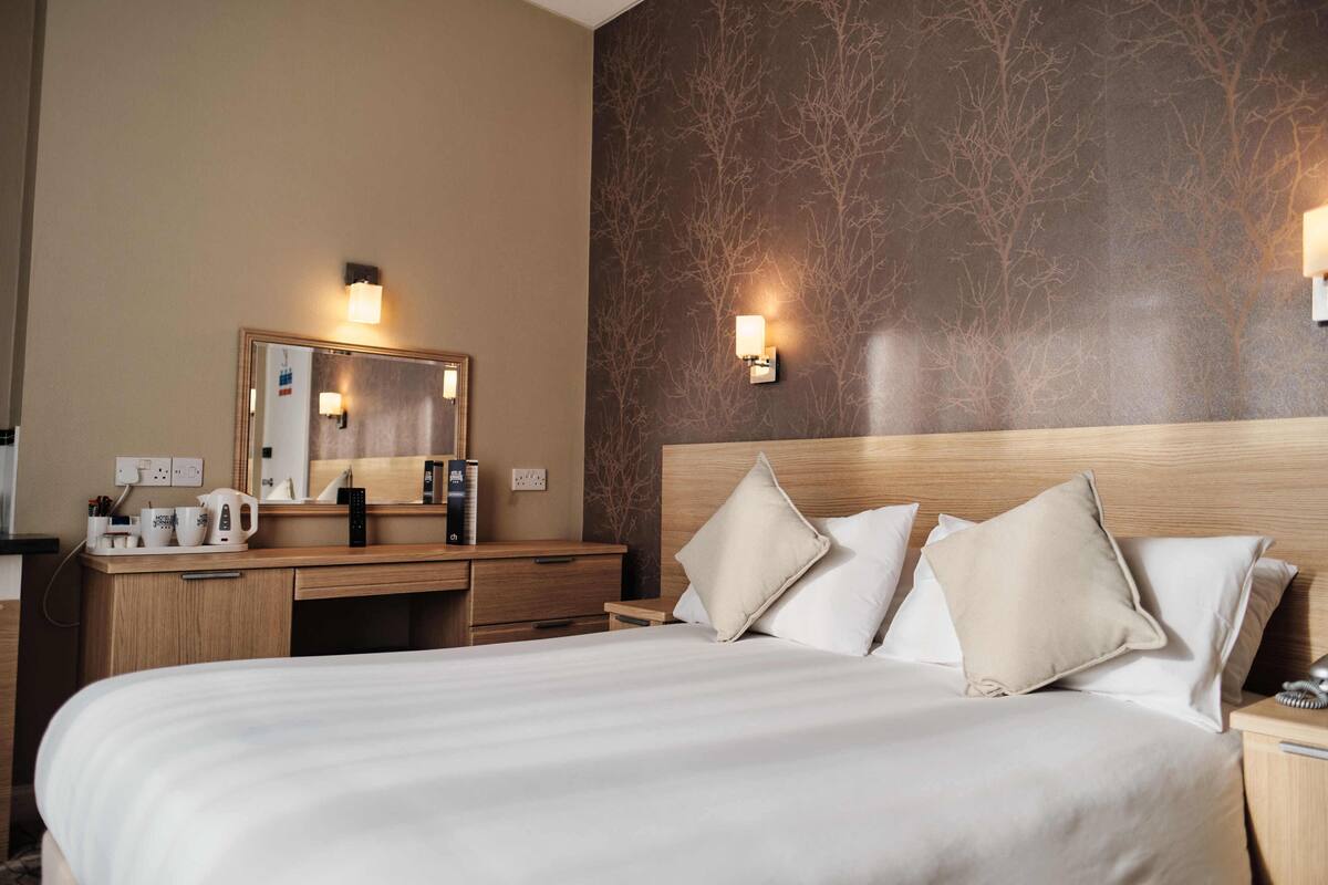 Habitación de hotel con una cama grande, una mesita de noche de madera, un escritorio con espejo y dos lámparas de pared.